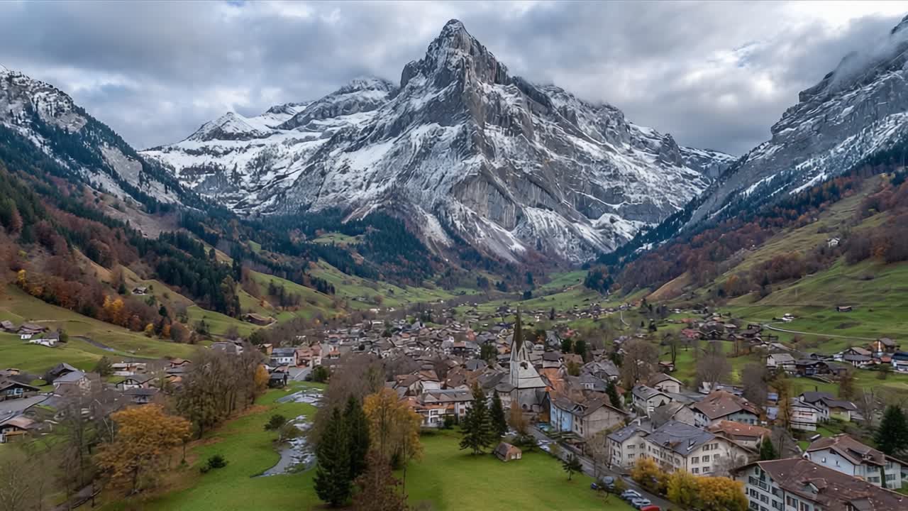 Splendida Vista Aerea di un Pittoresco Villaggio Alpino Incastonato in una Valle Rigogliosa e Circondato da Maestose Montagne Innevate Sotto un Cielo Drammatico