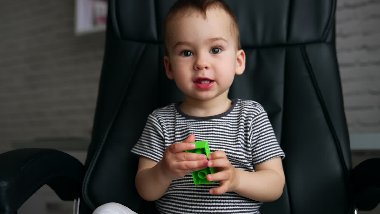 Beautiful Caucasian kid sits in a black office chair. Cute child turns a piece of constructor and says something.