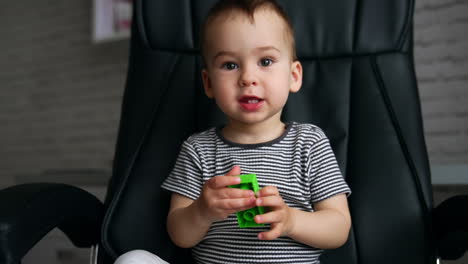 beautiful caucasian kid sits in a black office chair. cute child turns a piece of constructor and says something.