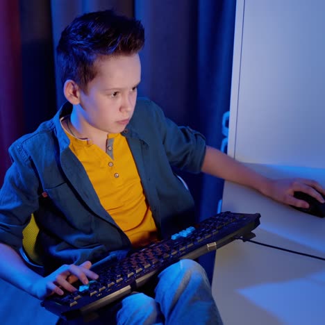 gamer playing games on a pc at home. boy is pushing buttons on a keyboard on the desk while playing games in his room. blue light.