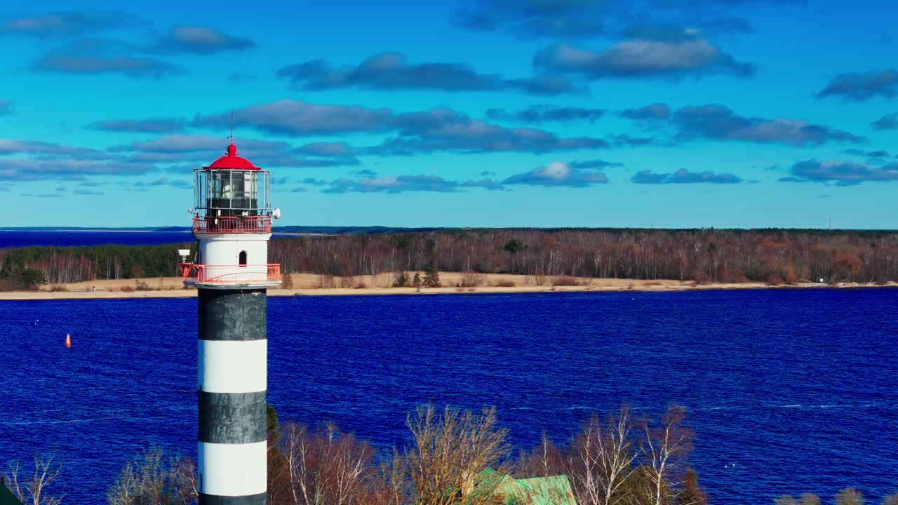 A tall black and white striped lighthouse with red top stands prominently above bare trees along a coastline. Deep blue water and a distant wooded shore complete the scenic landscape