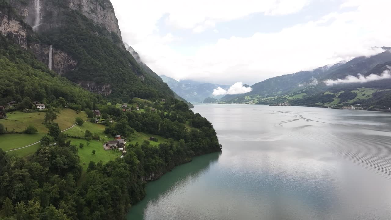 Drone view of Seerenbach Falls cascading into the Walensee in Switzerland. Scenic mix of lake, green meadows, forest, and towering alpine cliffs under cloudy skies