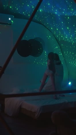 little girl plays and indulges with mother while jumping on bed in glamping bedroom. dimly lit room with bright projections of stars on white ceiling