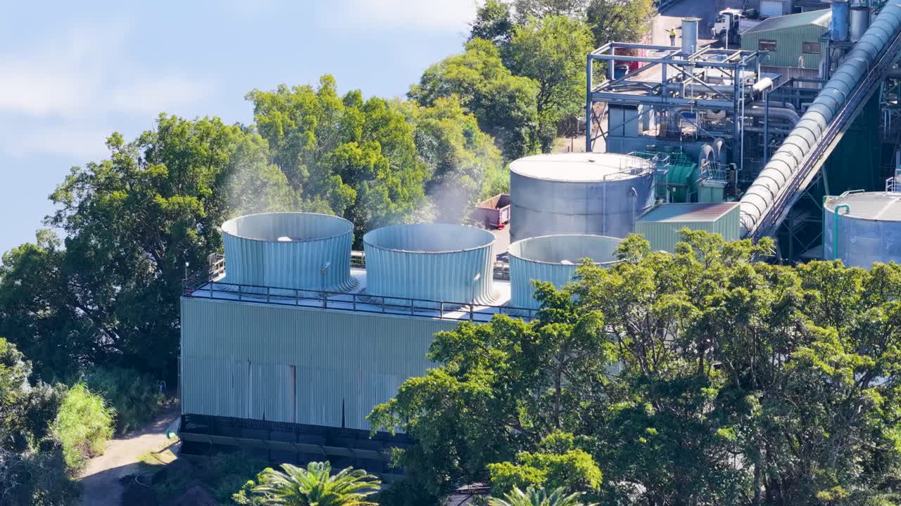 Cooling fans operate at an industrial facility surrounded by trees, emitting smoke under bright daylight