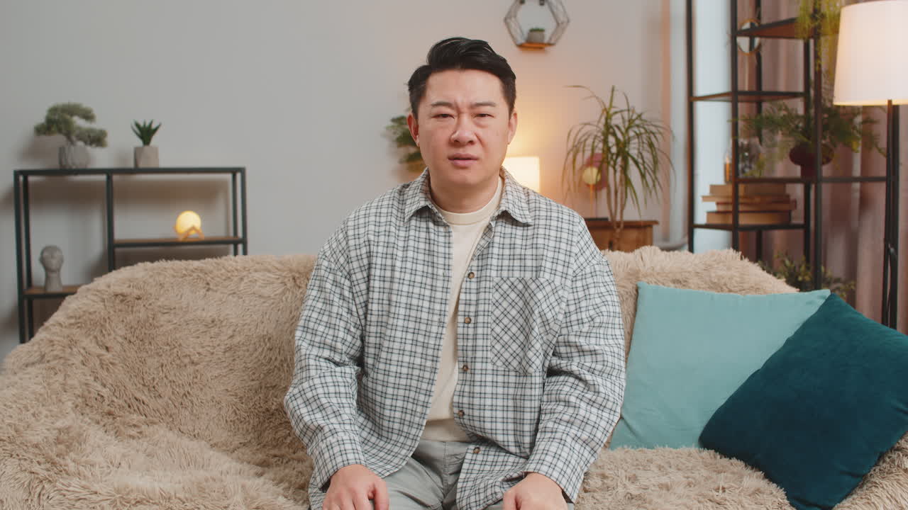 Confused asian handsome man feeling embarrassed about ambiguous question sitting on sofa at home