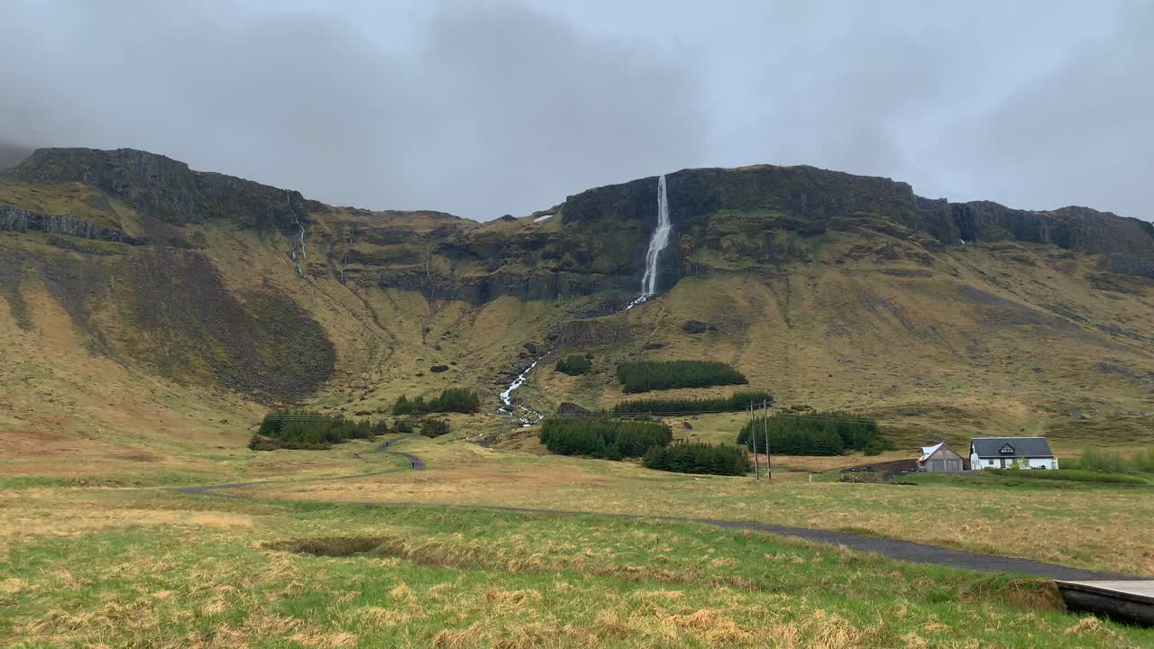 View over the high mountains around the tall waterfall Bjarnarfoss with a family home infront of it on a cloudy and misty day