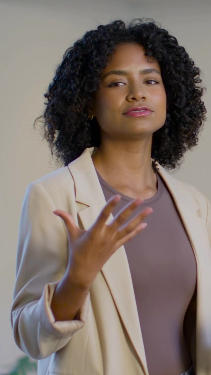 Vídeo vertical: Mulher fazendo gesto de pinça, abrindo a mão e falando em estúdio, blazer bege, planta à esquerda.