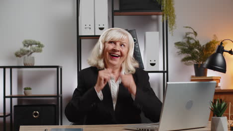cheerful senior businesswoman dancing and listening music through headphones at home office desk