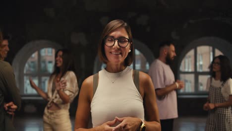 A-girl-psychologist-with-a-bob-hairstyle-in-glasses-and-a-white-T-shirt-poses-and-looks-at-the-camera-against-the-backdrop-of-her-group-therapy-in-a-brick-building