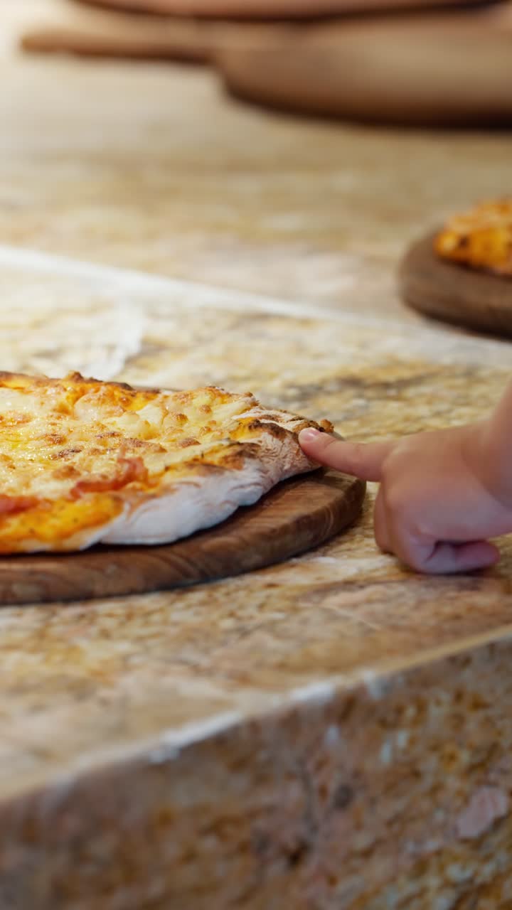 Vertical close-up shows a child's hand curiously touching the crust of a fresh pizza to check if it is hot during a cooking workshop