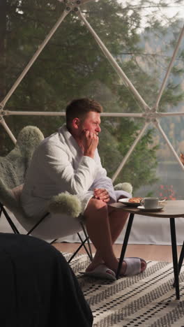couple enjoys morning meal in cottage. young man and woman in bathrobes drink coffee near panoramic window. magnificent mountain view during morning rituals