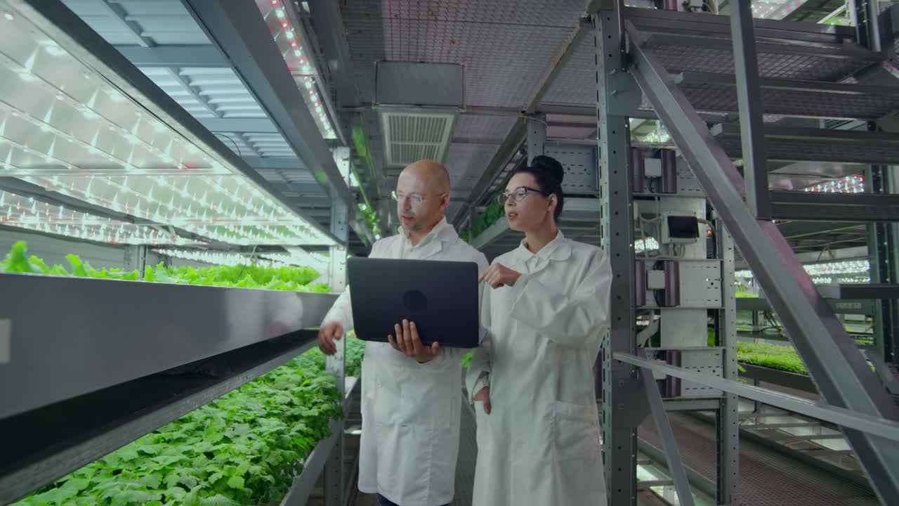 Scientists in white coats with a laptop and a tablet on a vertical farm with hydroponics make research data on vegetables in the data center for the analysis and programming of watering plants.