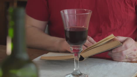 Man-reads-book-at-table-in-background-with-glass-of-red-wine