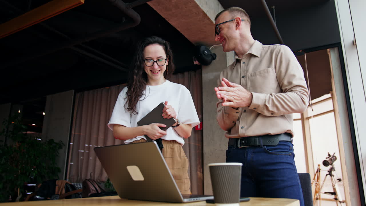 Cheerful office workers in the spacious office. Colleagues stand near the desk with laptop and discuss project. Low angle view.