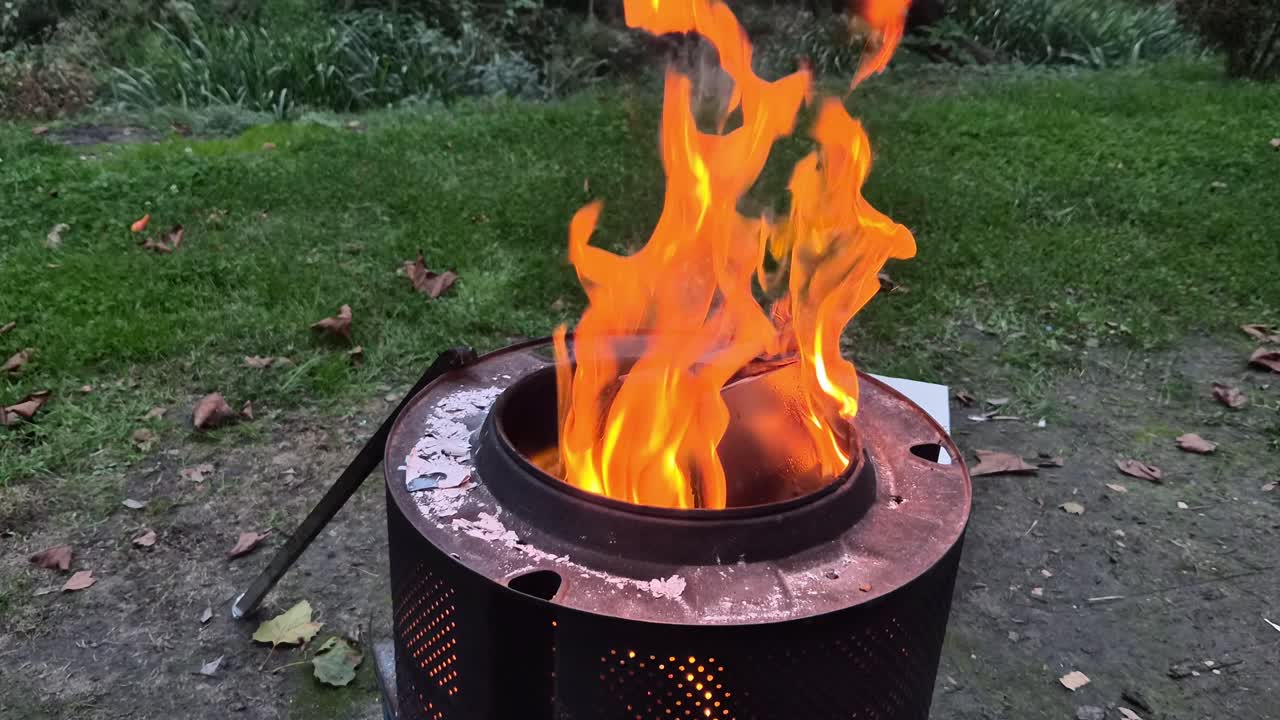 Flames rise high from a metal fire pit outdoors, glowing orange against the dark background