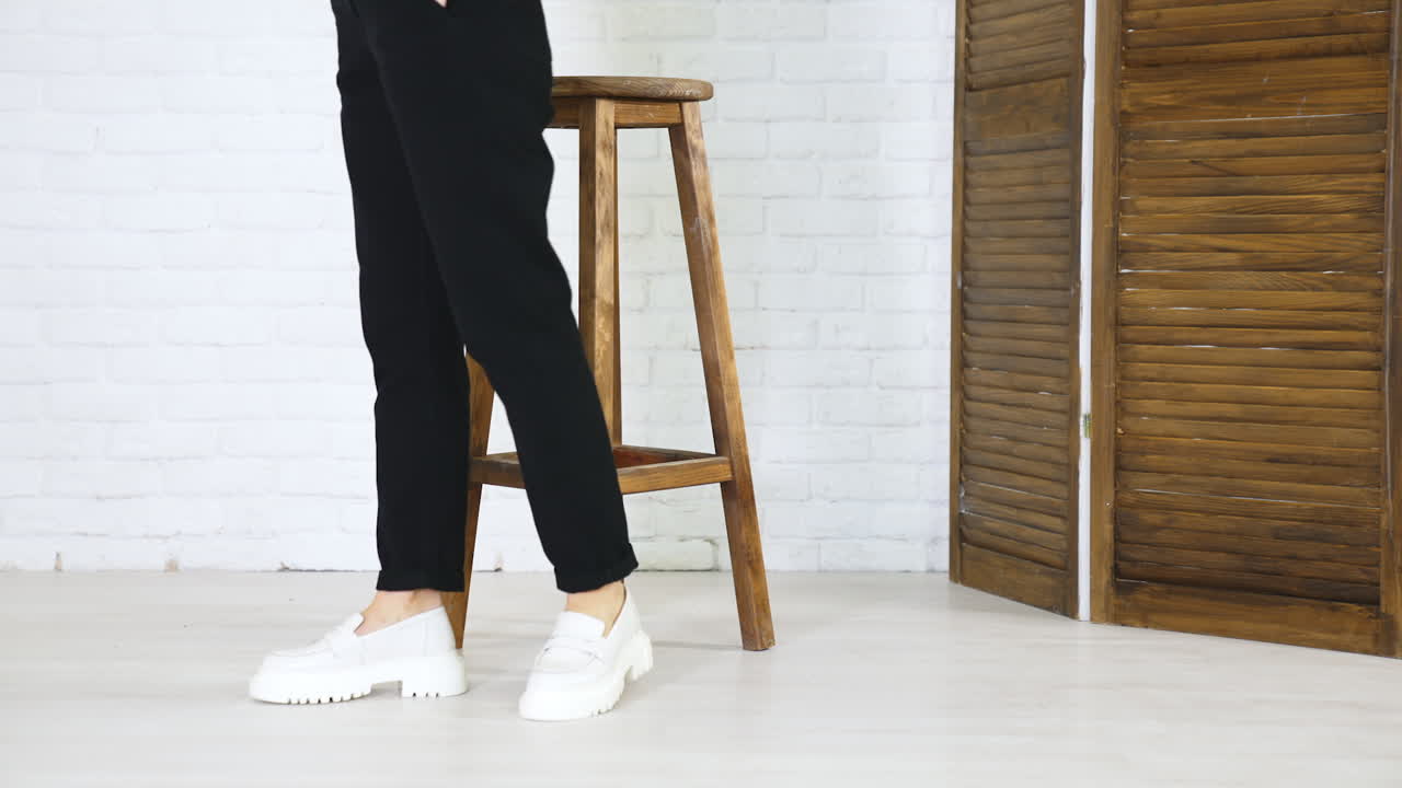 Female model posing in massive modern white shoes. Lady in black jeans walks in new pair of footwear in front of wooden stool. White wall backdrop.