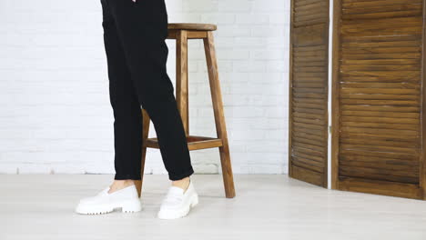 female model posing in massive modern white shoes. lady in black jeans walks in new pair of footwear in front of wooden stool. white wall backdrop.