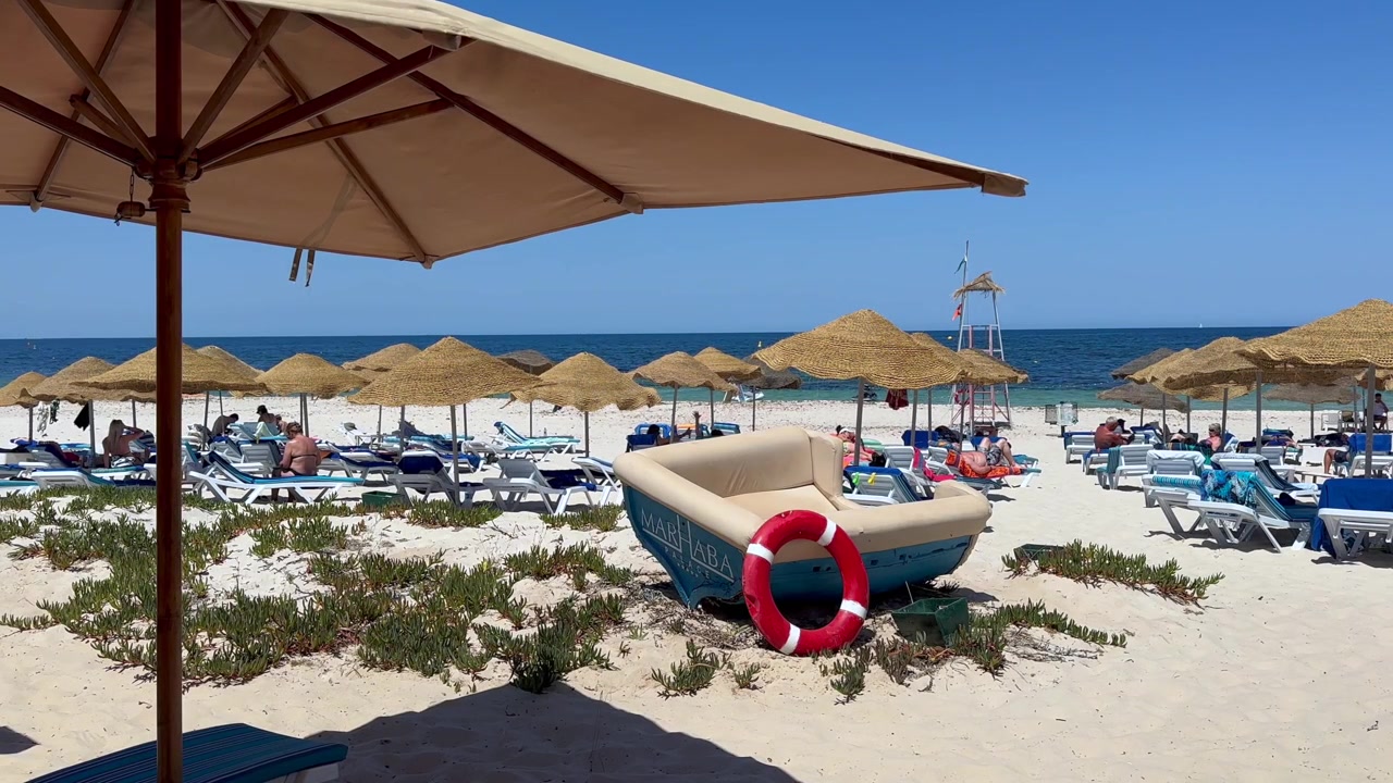 Vacanziere si rilassano su lettini sotto ombrelloni di paglia su una spiaggia sabbiosa mediterranea, con una piccola barca e un salvagente in primo piano in una limpida giornata estiva. Port El Kantaoui, Tunisia