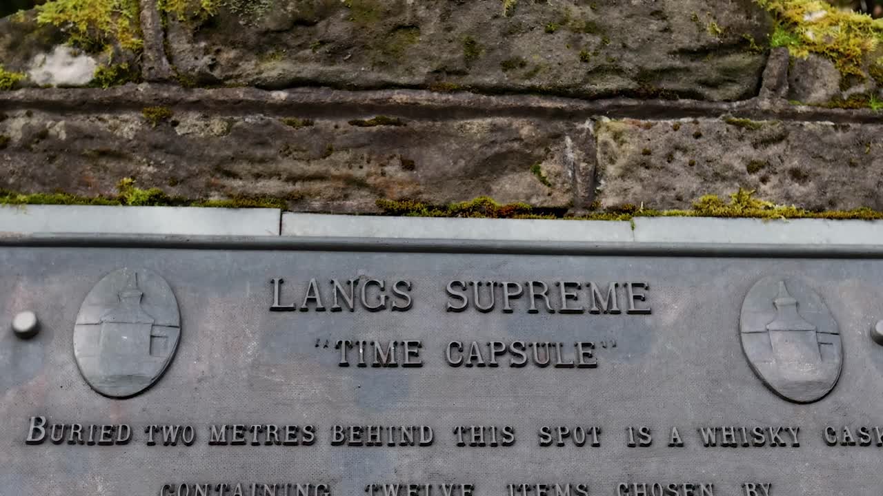 Close-up view of a metal plaque on a moss-covered stone wall, commemorating a time capsule buried in 1983.