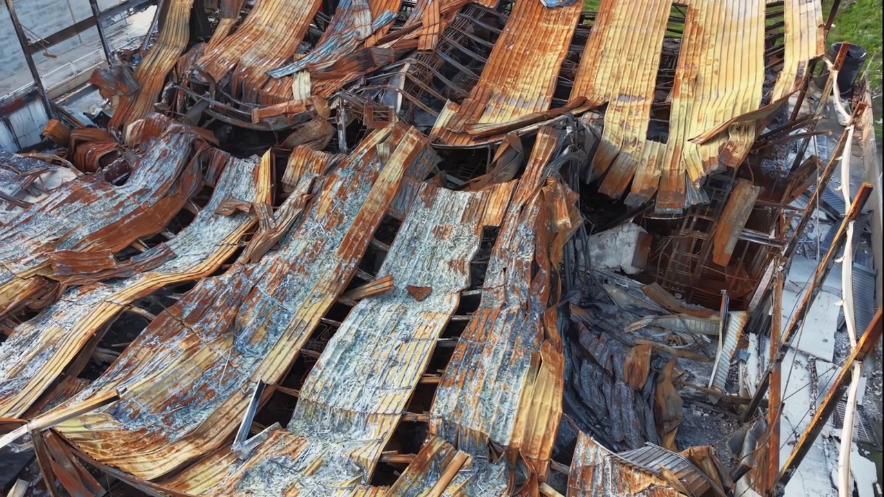 Collapsed roof structure of a building destroyed by fire, showing warped corrugated metal sheets, charred wooden beams and twisted steel framework after a devastating blaze