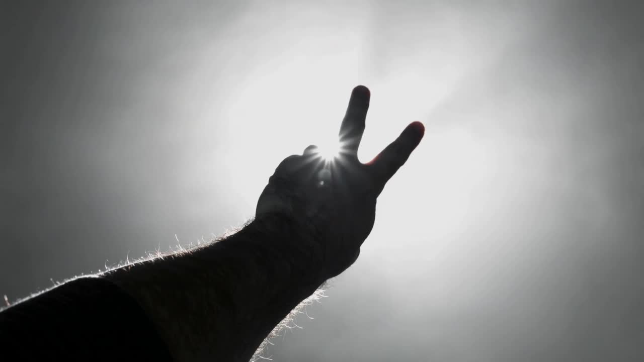 Reaching arm making V-sign toward sun in blue sky creating flare between fingers with ring tattoo