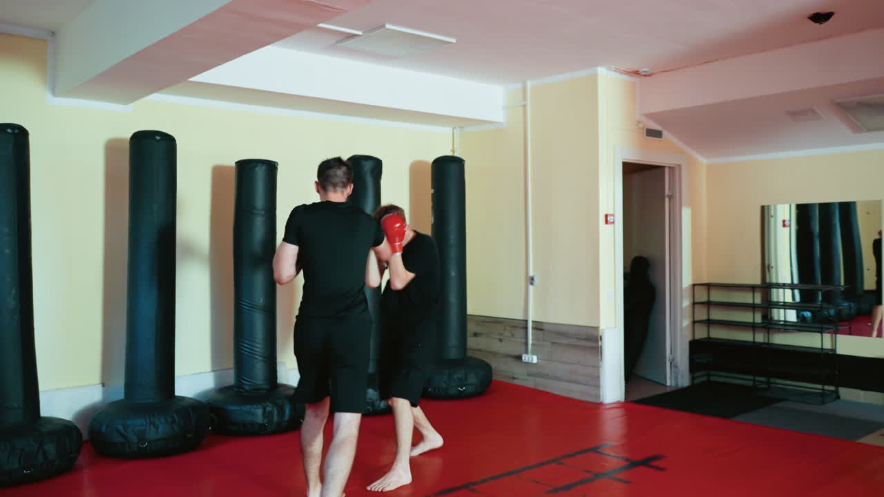 Fighters facing each other in combat stance inside gym, wearing boxing gloves and black outfits, preparing to spar on red mat floor surrounded by standing punching bags during martial arts training