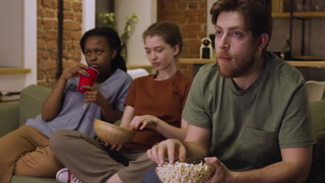 three roommates are watching a movie while eating popcorns sitting on sofa at home