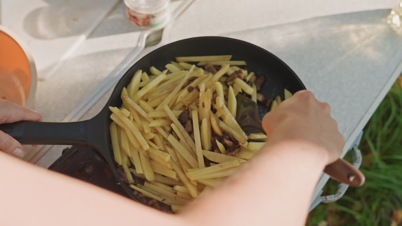 Camping skillet fries portable table caucasian woman stirring potatoes over camp stove, flipping golden fries with wooden spatula, adding seasoning, casual morning, grass background, nostalgic outdoor