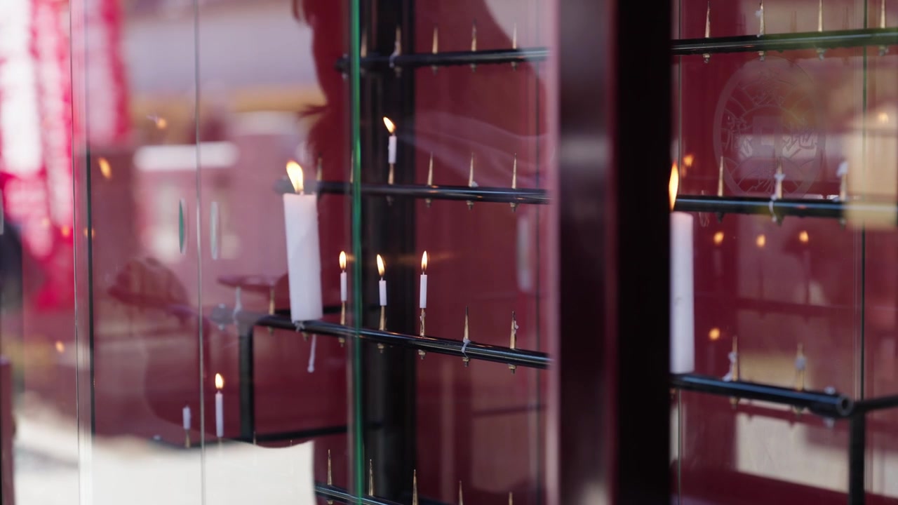 Pessoa com jaqueta escura em pé diante de fileiras de velas votivas brancas acesas dentro de um armário de santuário laqueado de vermelho em Tóquio.