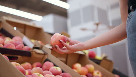 close-up of woman picking a red apple from a display, observing it with a focused gaze, while another shopper walks by in the background at a grocery store