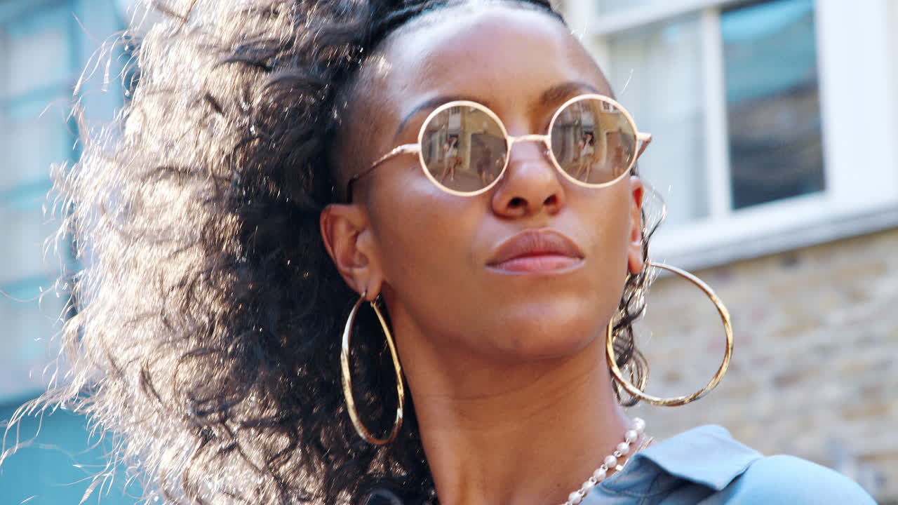 Fashionable young black woman wearing blue dress and sunglasses standing on the street, backlit, head and shoulders low angle