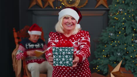 Senior-grandmother-in-New-Year-sweater-and-hat-presenting-Christmas-gift-box,-holidays-at-home