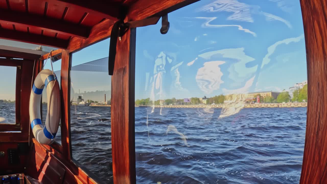 Cozy wooden sightseeing boat gliding along the Daugava River, showing Riga’s waterfront through a sunlit cabin window with reflections dancing across the water