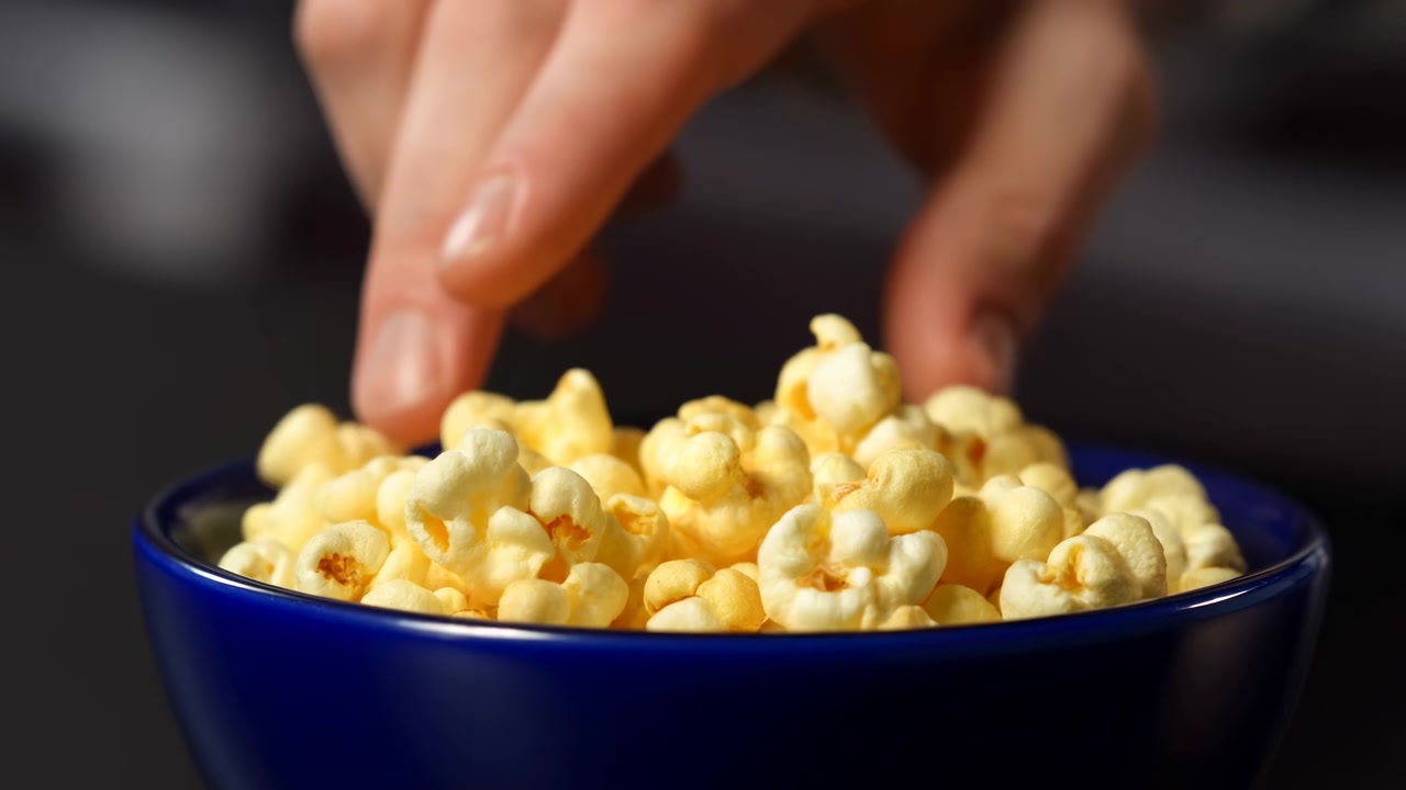 Dita si muovono verso il basso verso un mucchio di popcorn gialli all'interno di una ciotola blu lucida, con chicchi texturizzati illuminati vividamente in primo piano mentre l'ambiente scuro rimane leggermente sfocato