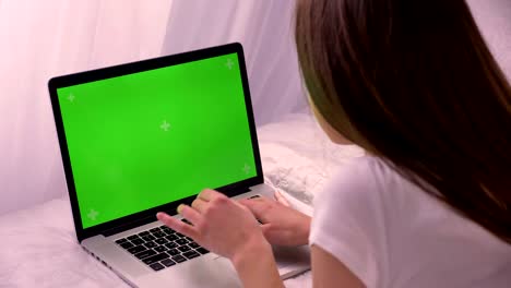 closeup back view of young attractive caucasian female typing on the laptop with green chroma-key screen lying on the bed at home indoors