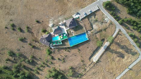 Aerial perspective showing several houses with a pool and sports court, located in an expansive, dry countryside area, surrounded by sparse vegetation and winding roads