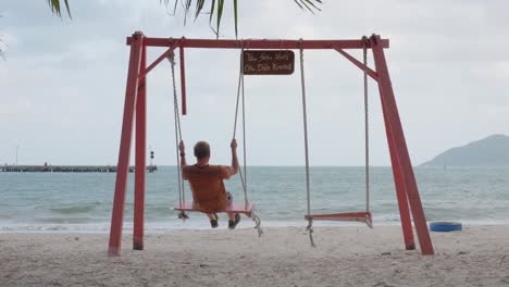 Hombre-Disfrutando-Del-Columpio-En-La-Playa-Mientras-Mira-Al-Océano