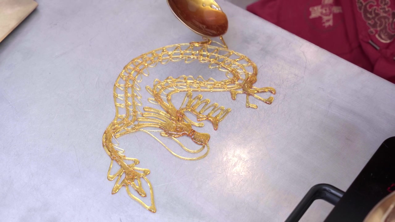 A near-complete tanghua sugar dragon glistens on a steel slab as the artisan adds final syrup threads with a bronze ladle, Thailand Chinese New Year fair. Part 4 of 6 — a tanghua sugar dragon created
