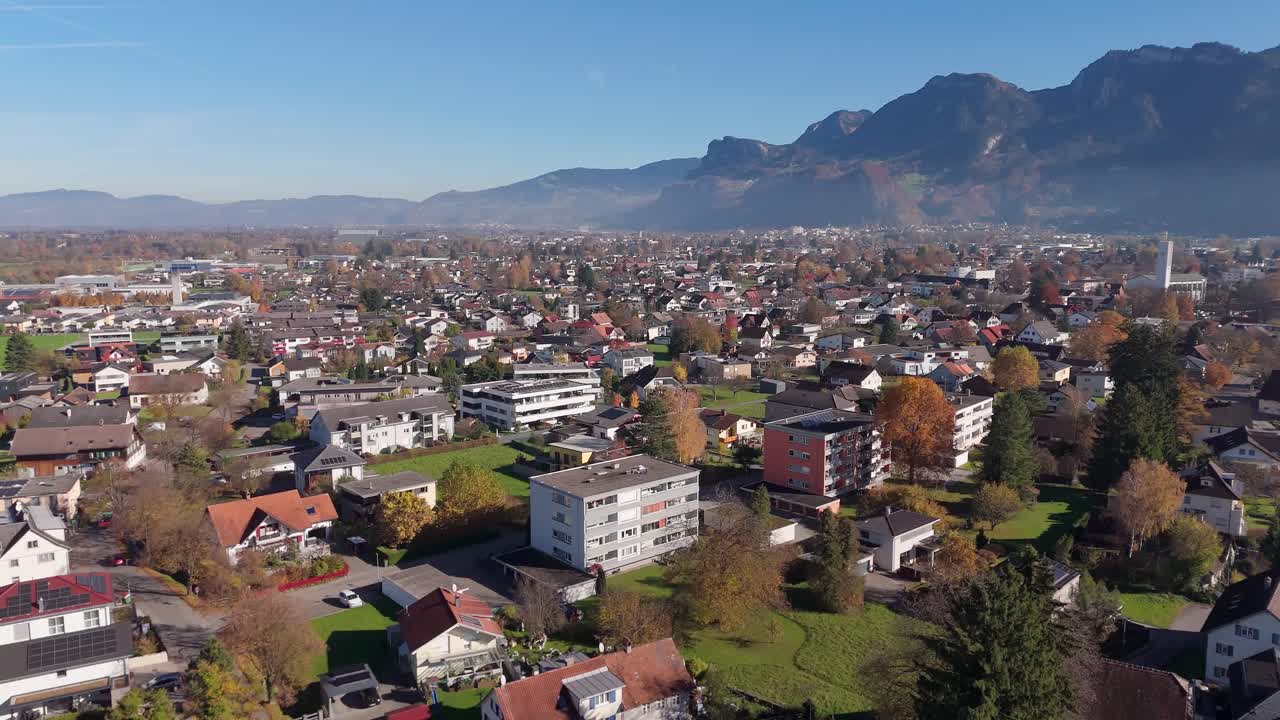 Aerial flyover austrian icy with apartment blocks and homes during sunny day. Fog hovering between alps mountains. Sunny day in fall season. Colored leaves of trees. Altach, Austria. Wide shot