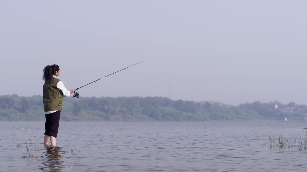 Seorang gadis Asia Selatan mengenakan rompi memancing berdiri sendirian di sebuah danau, melemparkan pancingnya ke air terbuka dalam keheningan