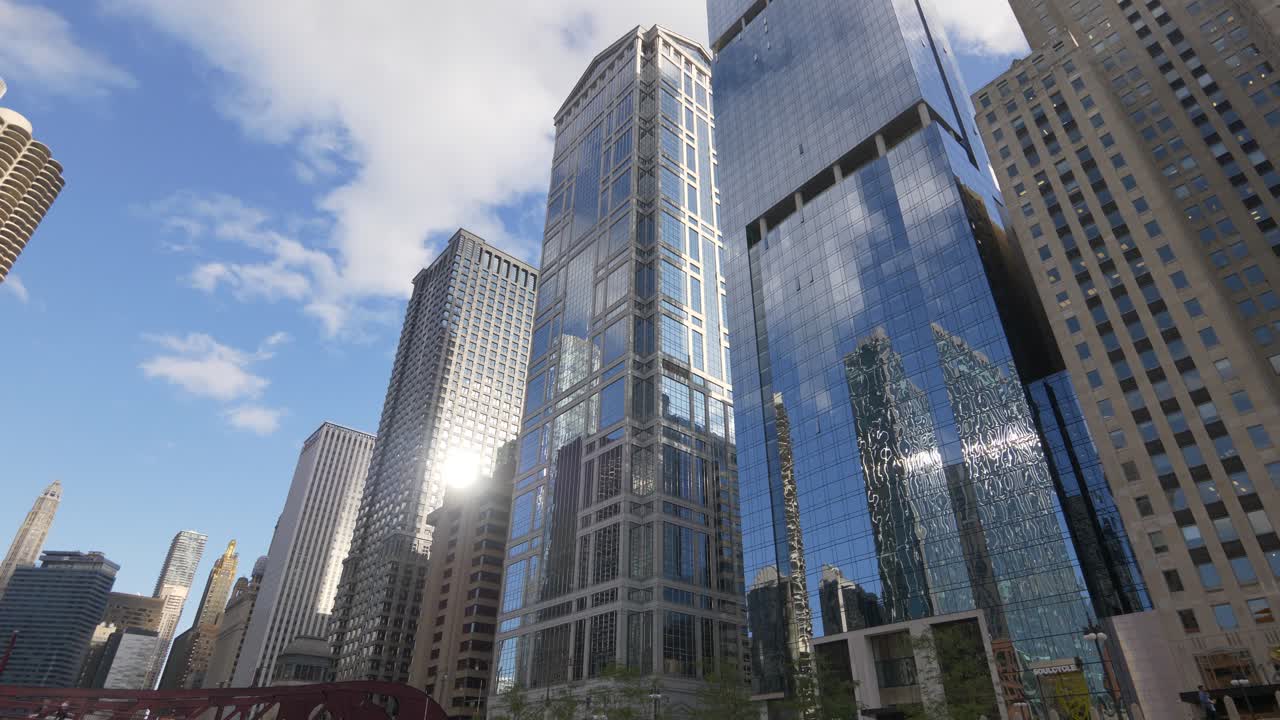 Stunning aerial footage of the Chicago skyline, showcasing towering skyscrapers reflecting sunlight against a bright, partly cloudy sky. Captures the bustling urban landscape from above