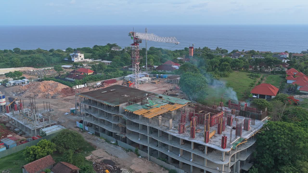 Pemandangan sudut tinggi yang memperlihatkan bangunan beton besar yang sedang dibangun di dekat samudra di lokasi tropis, dengan menara derek di dekatnya