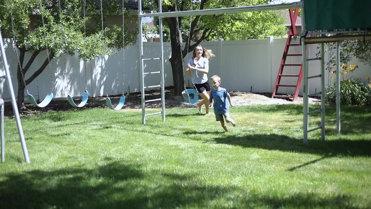 Slow Motion shot of a girl playfully chasing her younger brother around in the backyard by the playground.