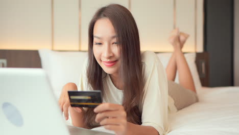 attractive asian woman lying on king size bed and using her credit card for shopping online, type in card information on the mobile phone and winning prize lottery, exited face expression, close-up