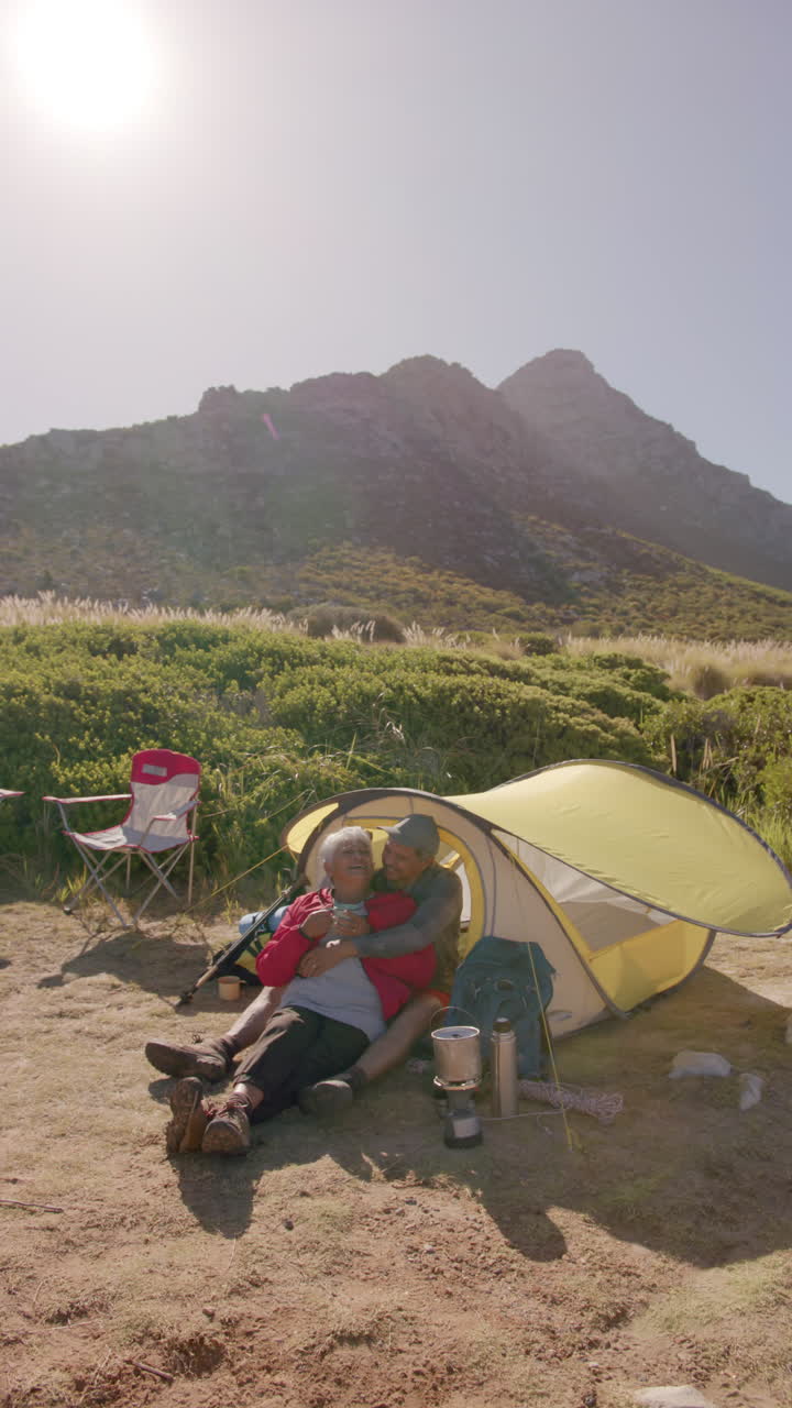 Vertical video of senior biracial couple embracing at tent in mountains, in slow motion