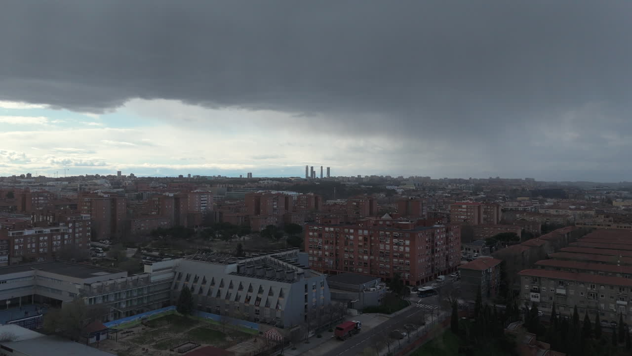 Stunning aerial view of Madrid with its iconic four towers in the distance. The cityscape is covered by dramatic storm clouds, contrasting with the urban skyline, streets, and residential buildings