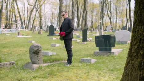 hombre de traje negro sosteniendo rosas rojas de pie frente a una tumba en un cementerio 1