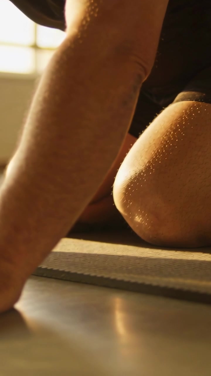 Video verticale: Uomo che si china appoggiando le mani sul tappetino vicino a una grande finestra, che si stira indossando dei pantaloncini.