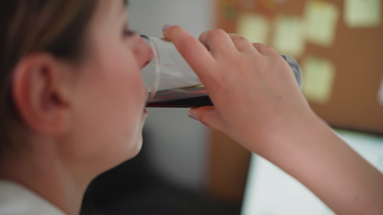 Rear view of woman drinking dark beverage from glass in home office setting, blurred sticky notes on wall and laptop screen visible in background