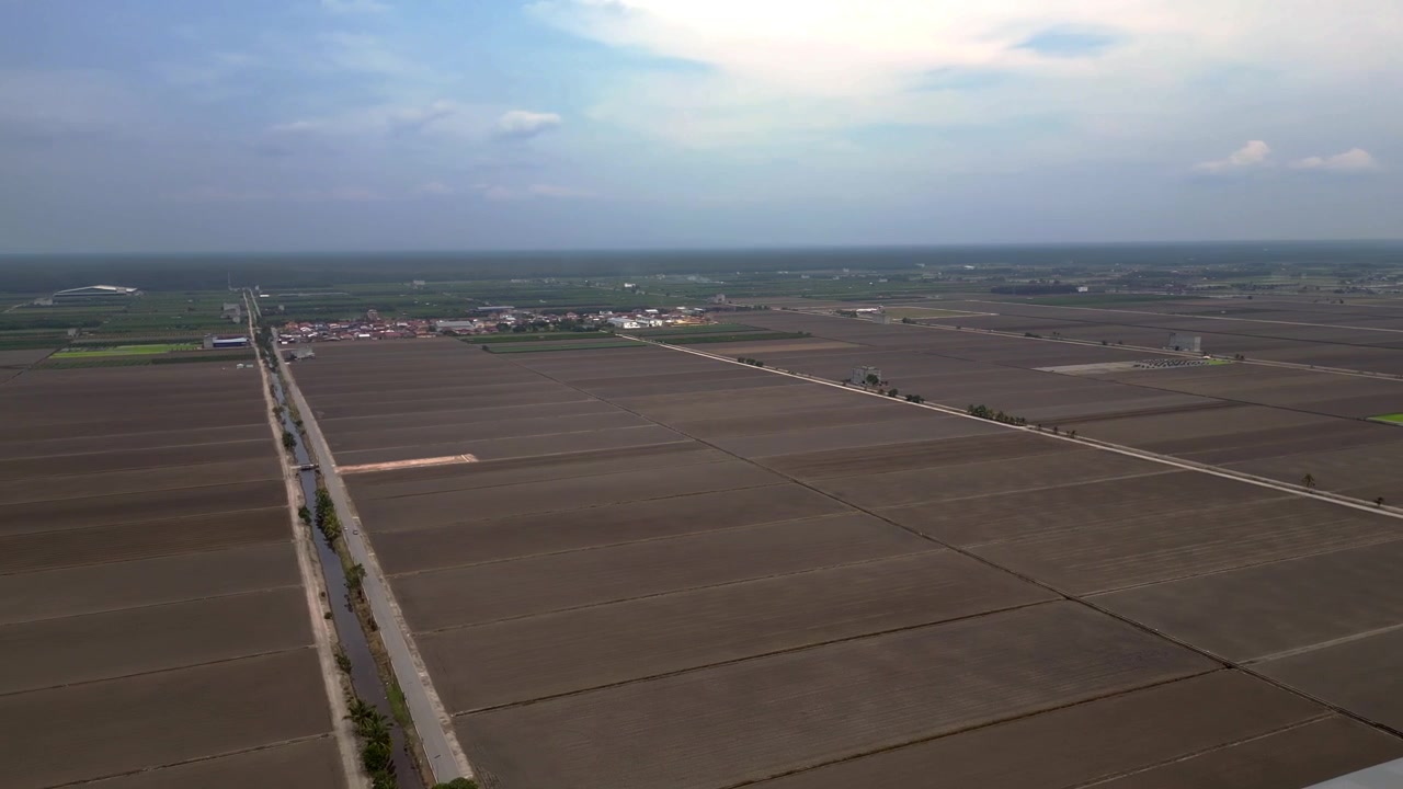 Pemandangan udara yang luas dari sawah Sekinchan Infinity yang telah dipanen, menampilkan petak-petak persegi panjang yang tandus, jalan setapak, dan sebuah bangunan di kejauhan, bersiap untuk budidaya. Pemandangan udara indah dengan putaran pan ke kanan